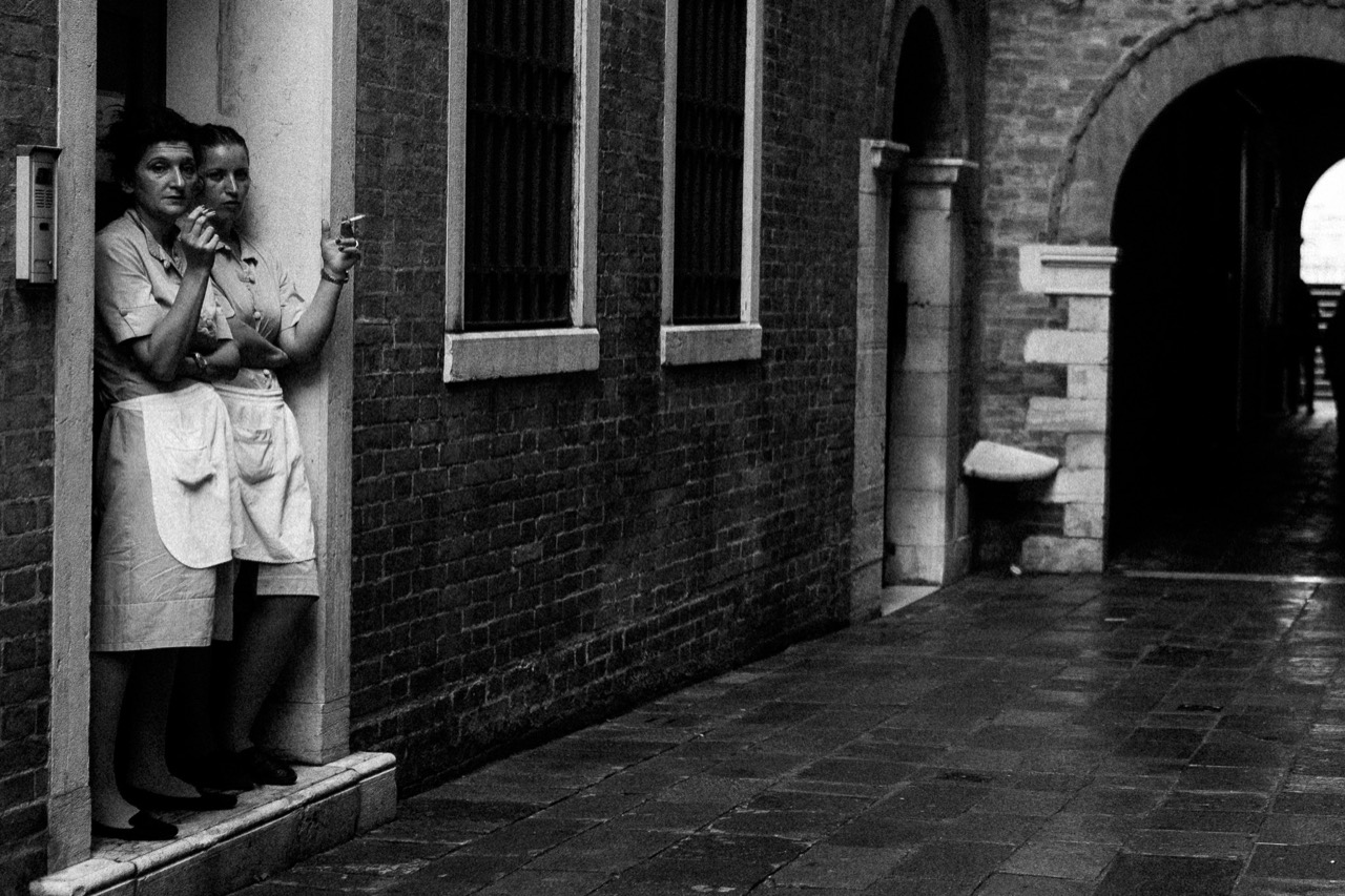A person standing beside a brick doorway, photographed in black and white